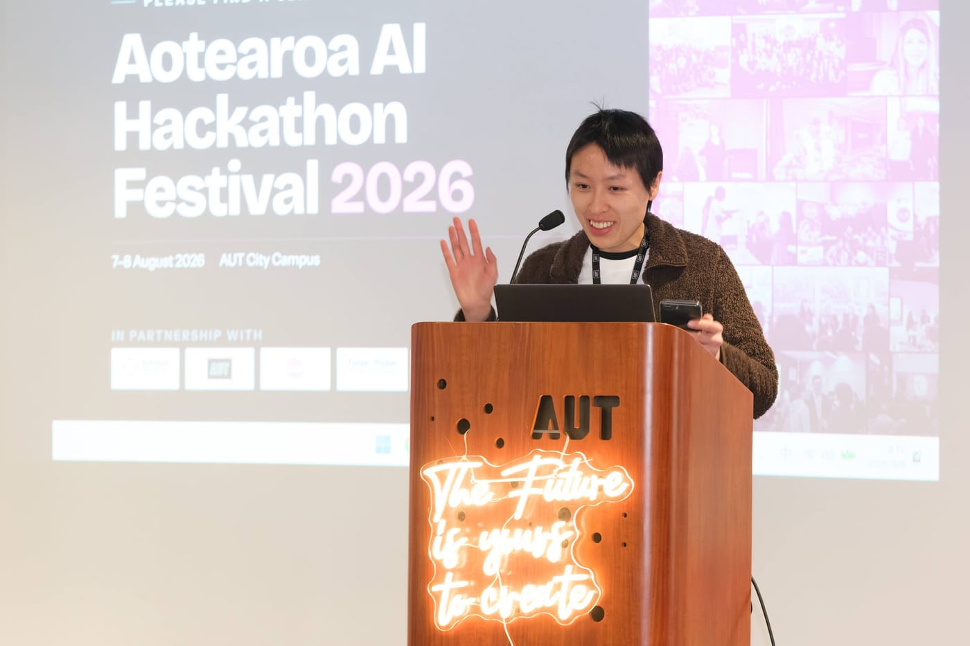 Chan Meng MCing the opening of the Aotearoa AI Hackathon Festival 2026 from the AUT podium, the festival title slide projected behind her